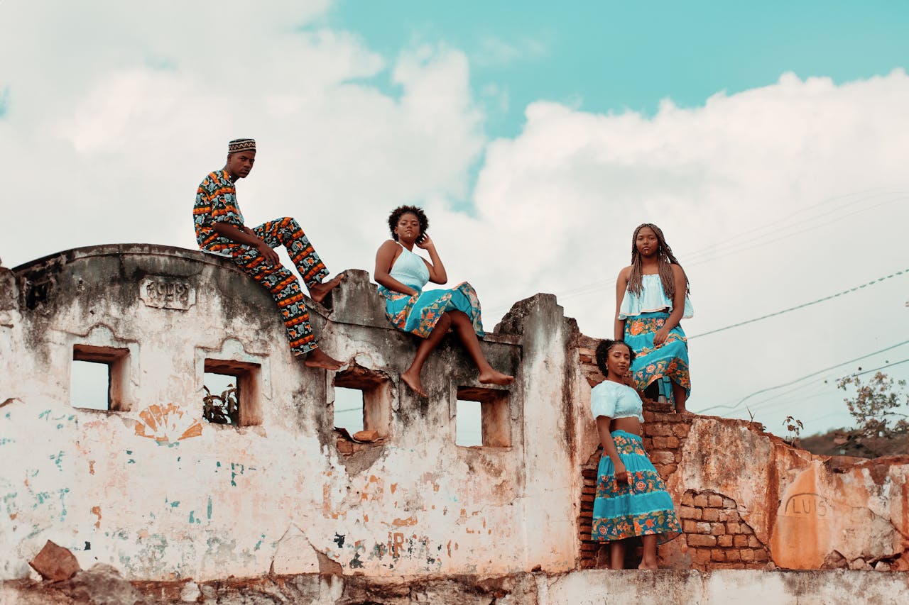 Four models wearing vibrant traditional African clothing pose on a rustic wall under a cloudy sky.