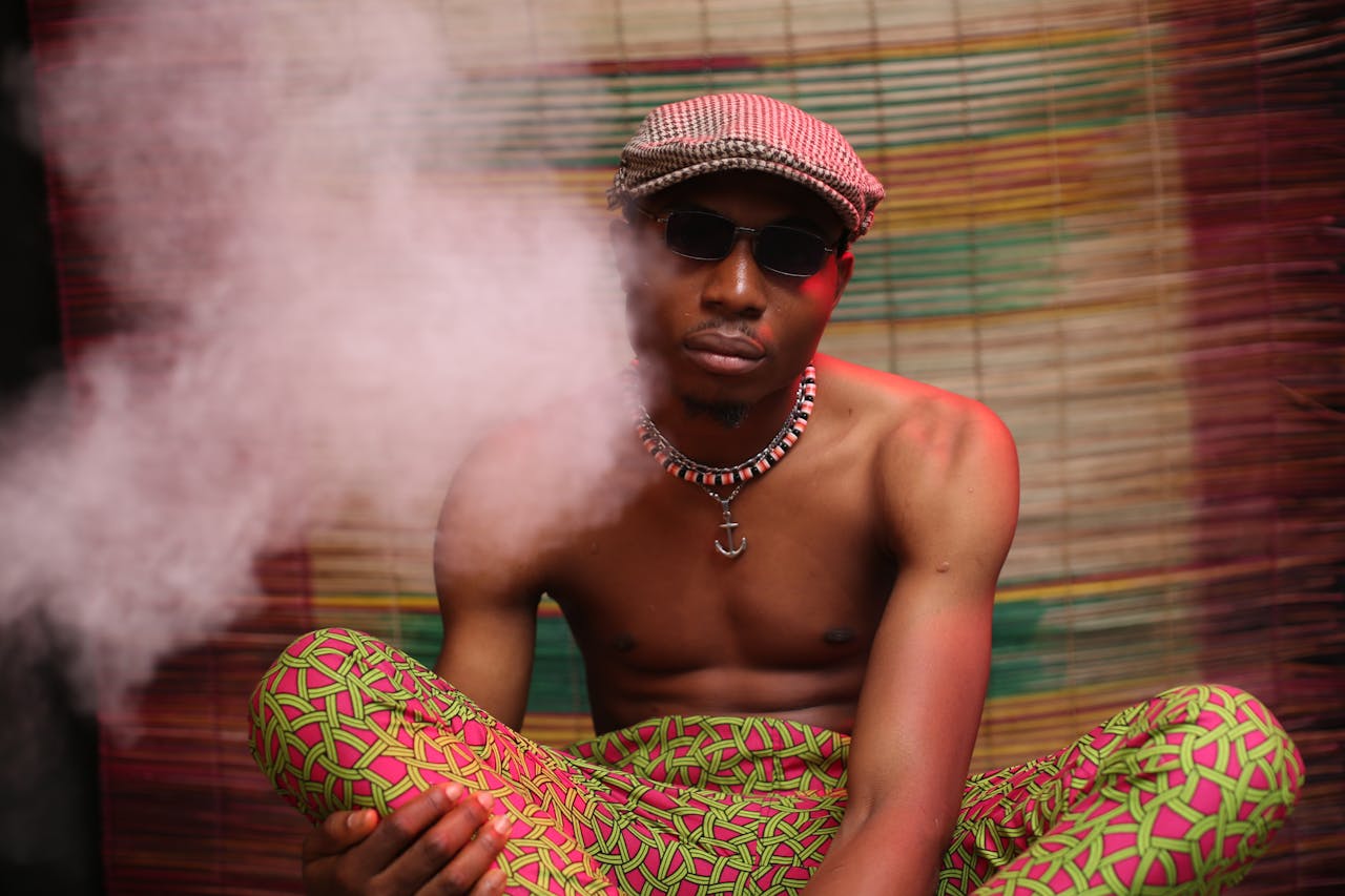 Shirtless black guy in colorful pants and cap sitting in dark room and smoking cannabis