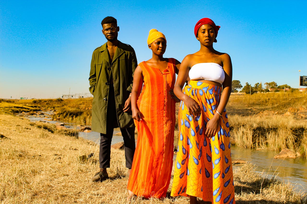 Three adults pose in traditional clothing in the outdoor setting of Soweto, South Africa, showcasing cultural attire and vibrant colors.